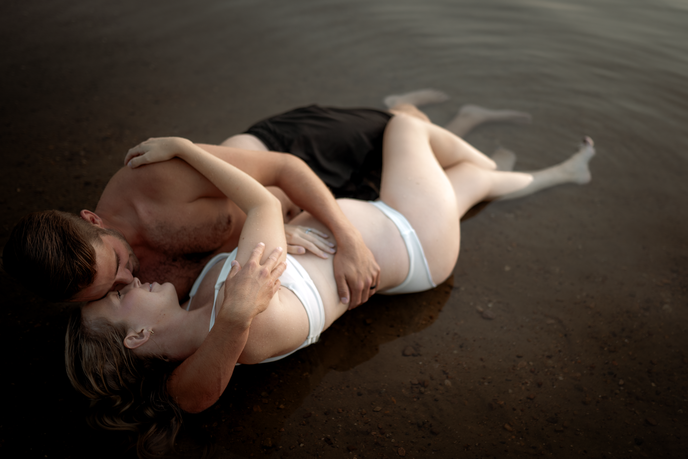 Pregnant couple laying on the lake shore embracing each other.